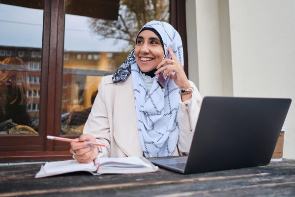 Young smiling Arabic woman in hijab happily talking on smartphone working on laptop on street